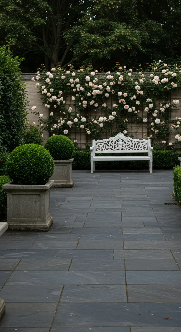 Terras met donkere tegels, witte tuinbank en een muur begroeid met lichtroze klimrozen, geflankeerd door Buxus.