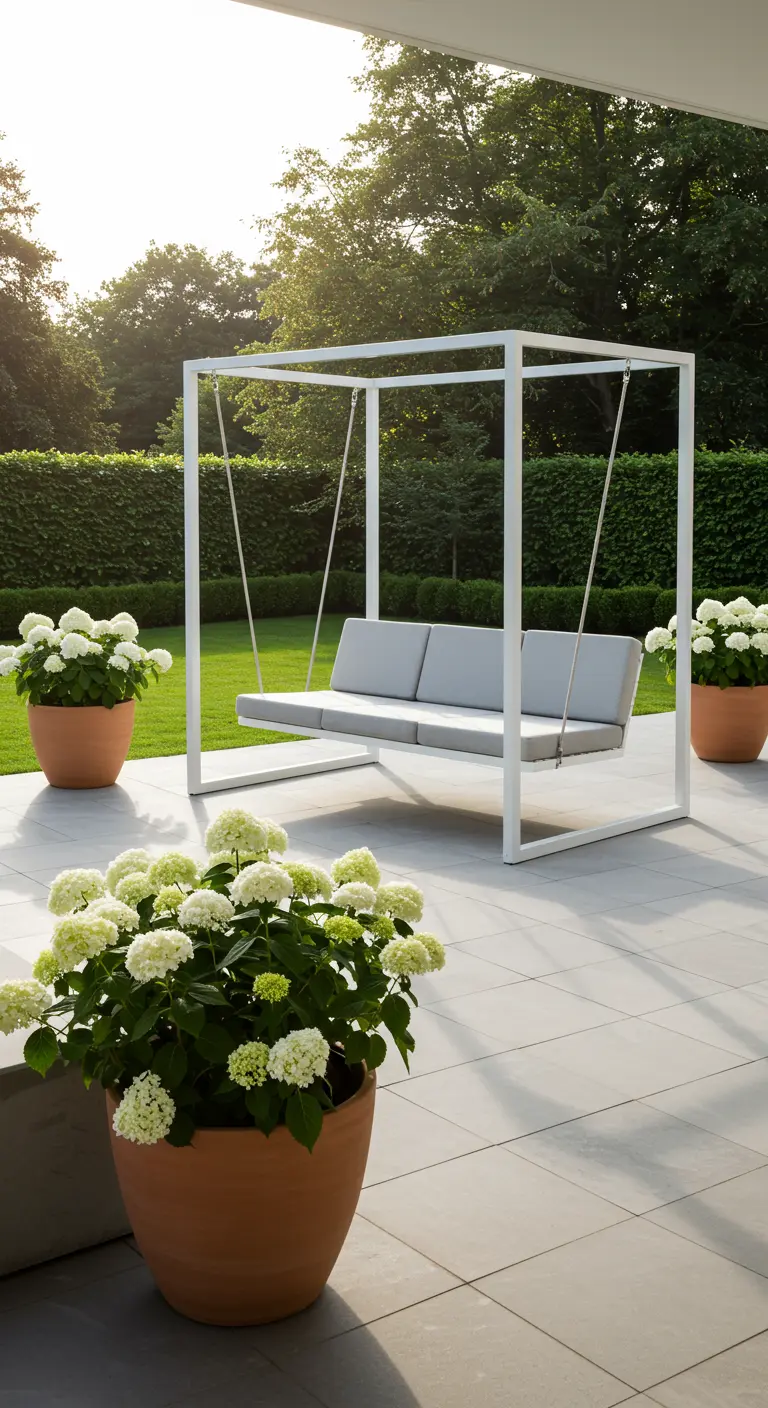 Witte metalen schommelbank met kussens op een terras, omringd door potten met witte bloemen.