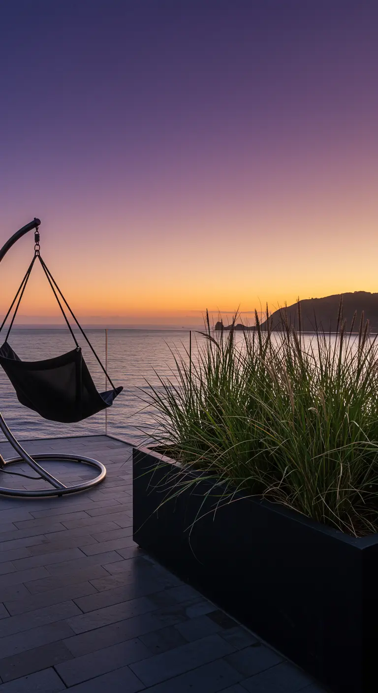 Zwarte hangstoel op terras met zeezicht bij zonsondergang en plantenbak met hoge grassen.