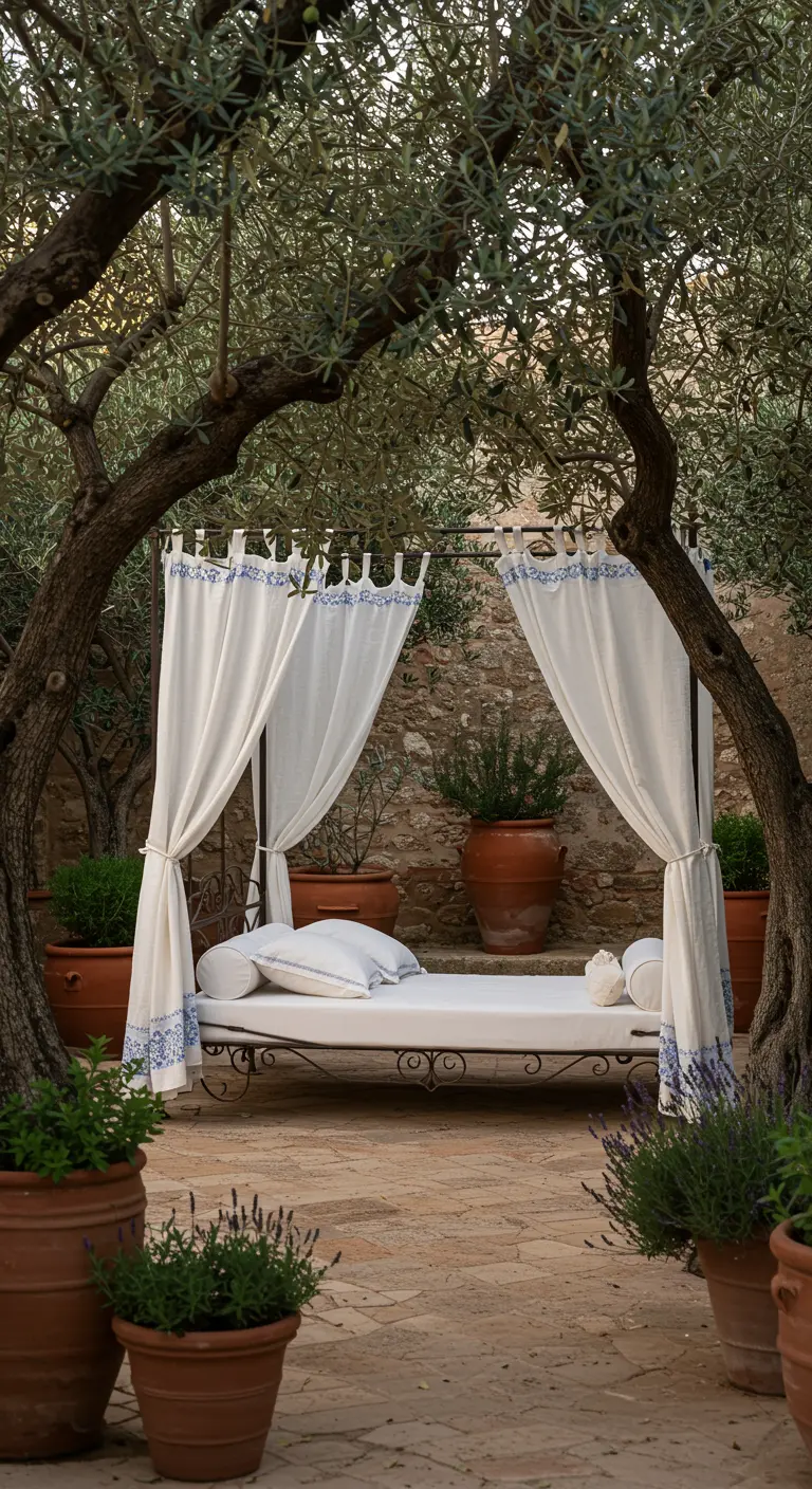 Zwevend smeedijzeren bed met witte en blauwe gordijnen tussen olijfbomen en terracotta potten in een Mediterrane tuin.